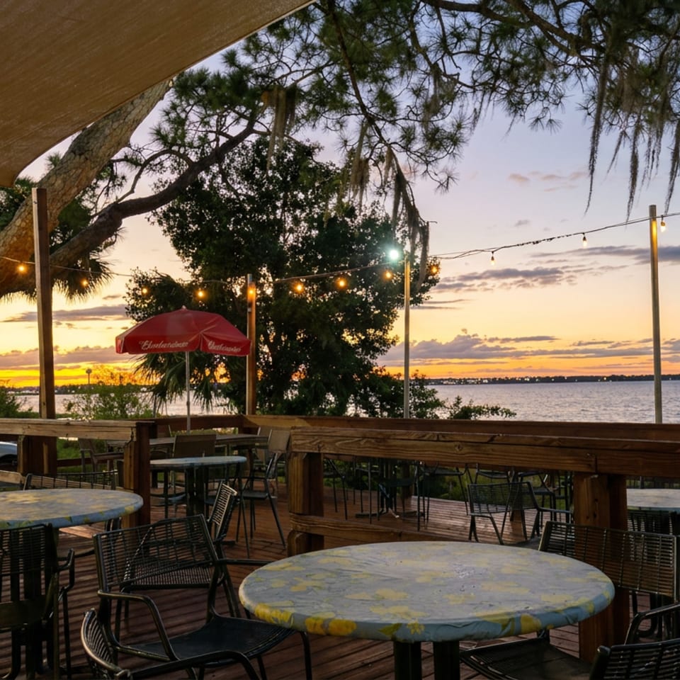 Outdoor restaurant terrace overlooking the water at sunset, with string lights, shaded tables, and a red umbrella.