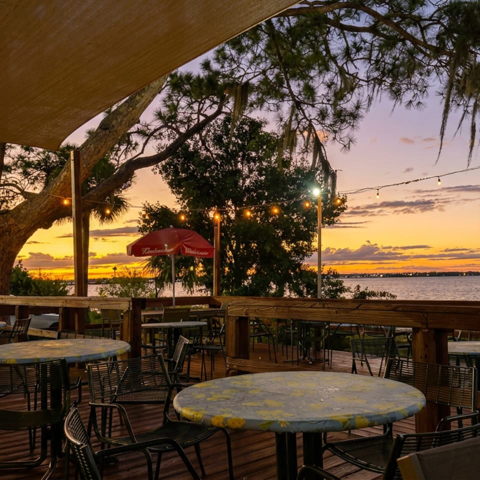 Outdoor restaurant deck with empty tables, string lights, waterfront views, and sunset beneath sprawling trees.