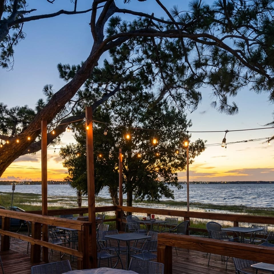Waterfront restaurant patio with outdoor tables, string lights, large pine trees, and a sunset over the bay.