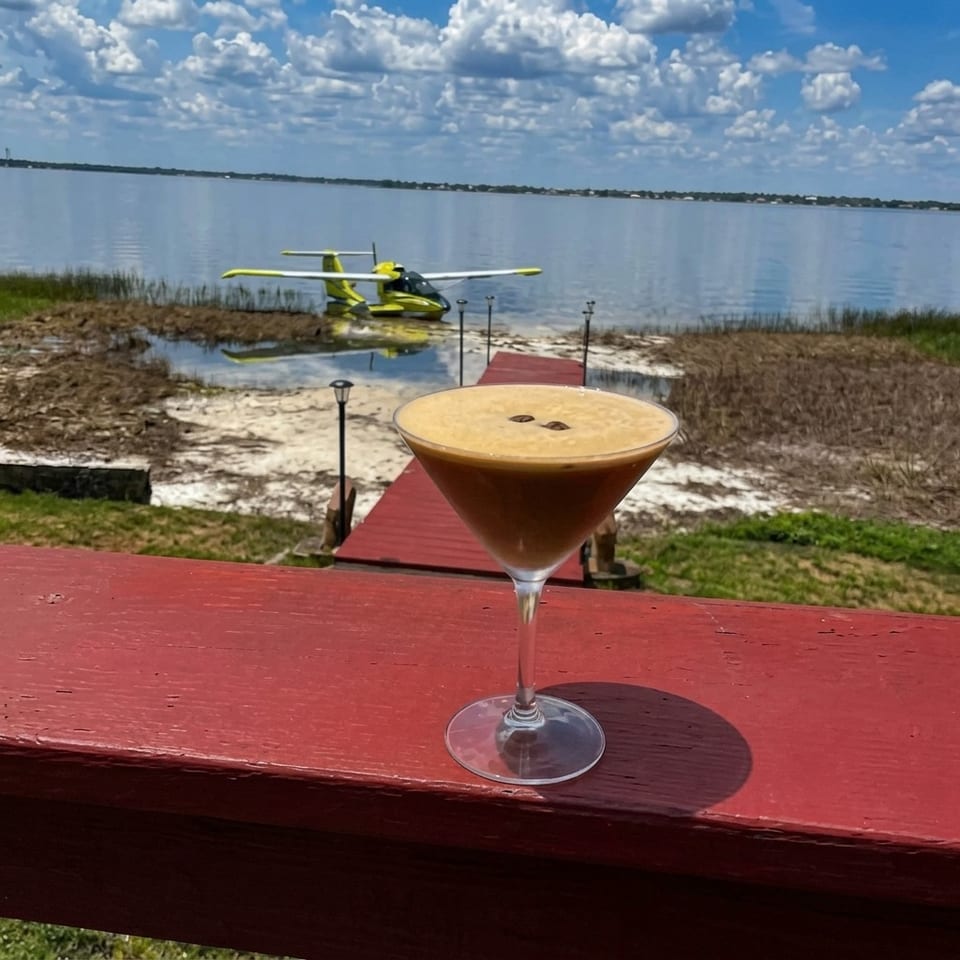 Foamy cocktail on a red wooden table overlooking a calm lake, dock, and parked seaplane beneath a partly cloudy sky.