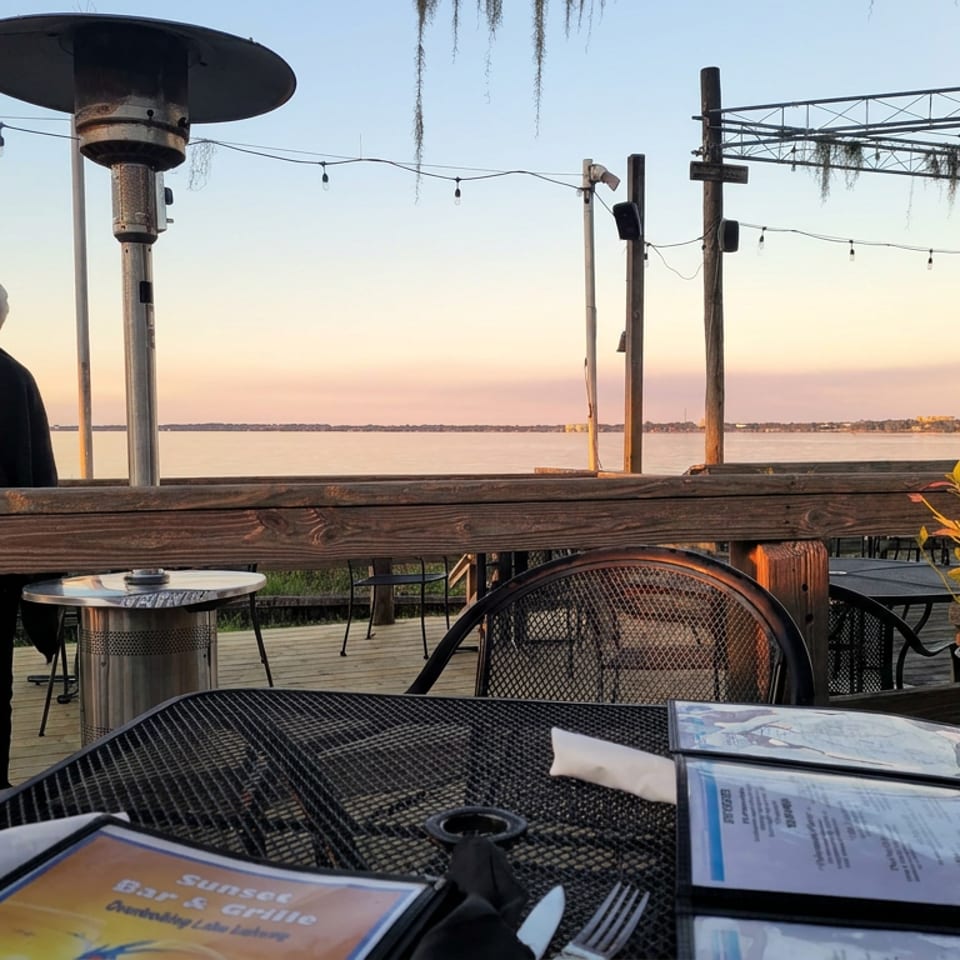 Waterfront restaurant patio with menus, outdoor heaters, string lights, and a person overlooking calm water at sunset.