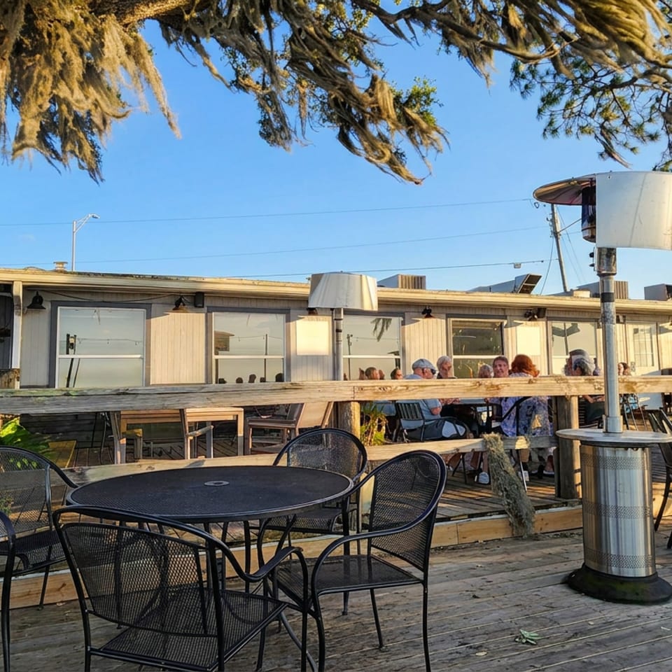 Outdoor restaurant patio with black metal tables, chairs, heaters, wooden decking, diners, and oak trees under a clear blue sky.