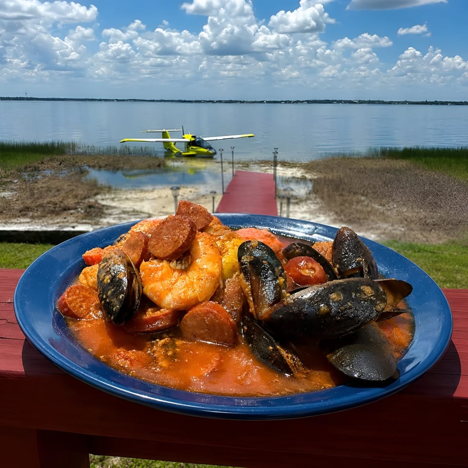 Seafood stew with shrimp, mussels, clams, and sausage on a blue plate overlooking a calm bay and seaplane.