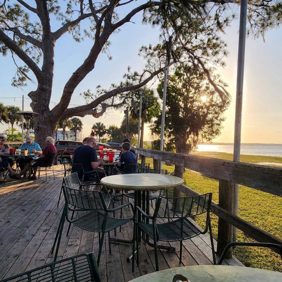Waterfront restaurant deck with outdoor tables, diners, palm trees, and sunset views across the calm bay.