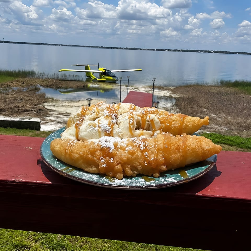 Plate of powdered sugar and caramel-drizzled fried pastries on a waterfront table, with a seaplane in the background.