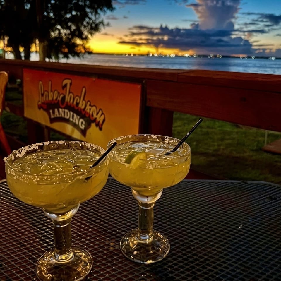 Two salted-rim margaritas sit on a restaurant patio table overlooking the ocean at sunset.