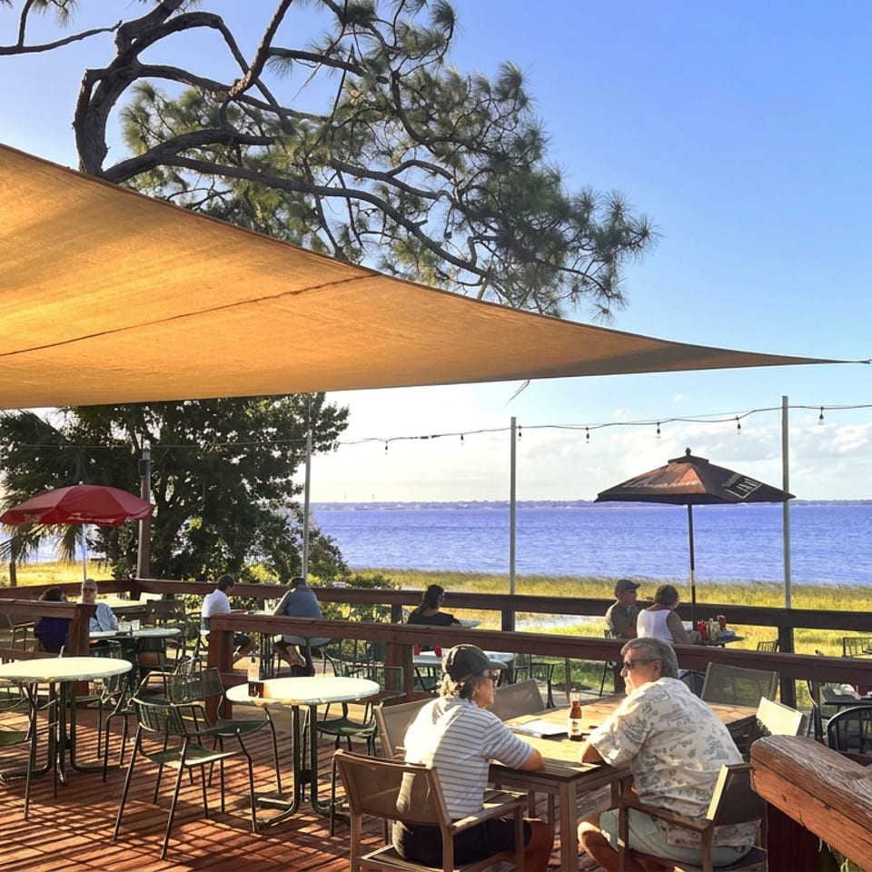 Outdoor waterfront restaurant patio with diners, string lights, umbrellas, and ocean views beneath a sunny sky.