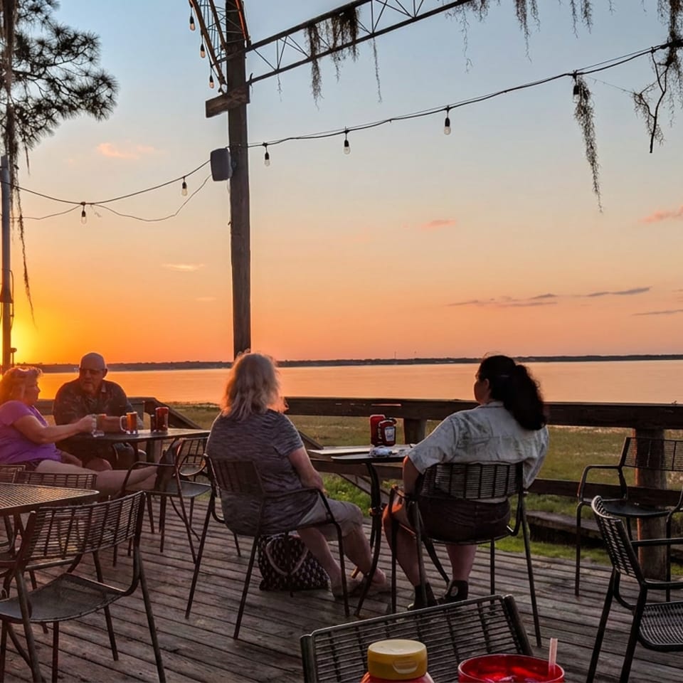 Outdoor waterfront restaurant patio with diners, string lights, metal tables, and a glowing sunset over calm water.