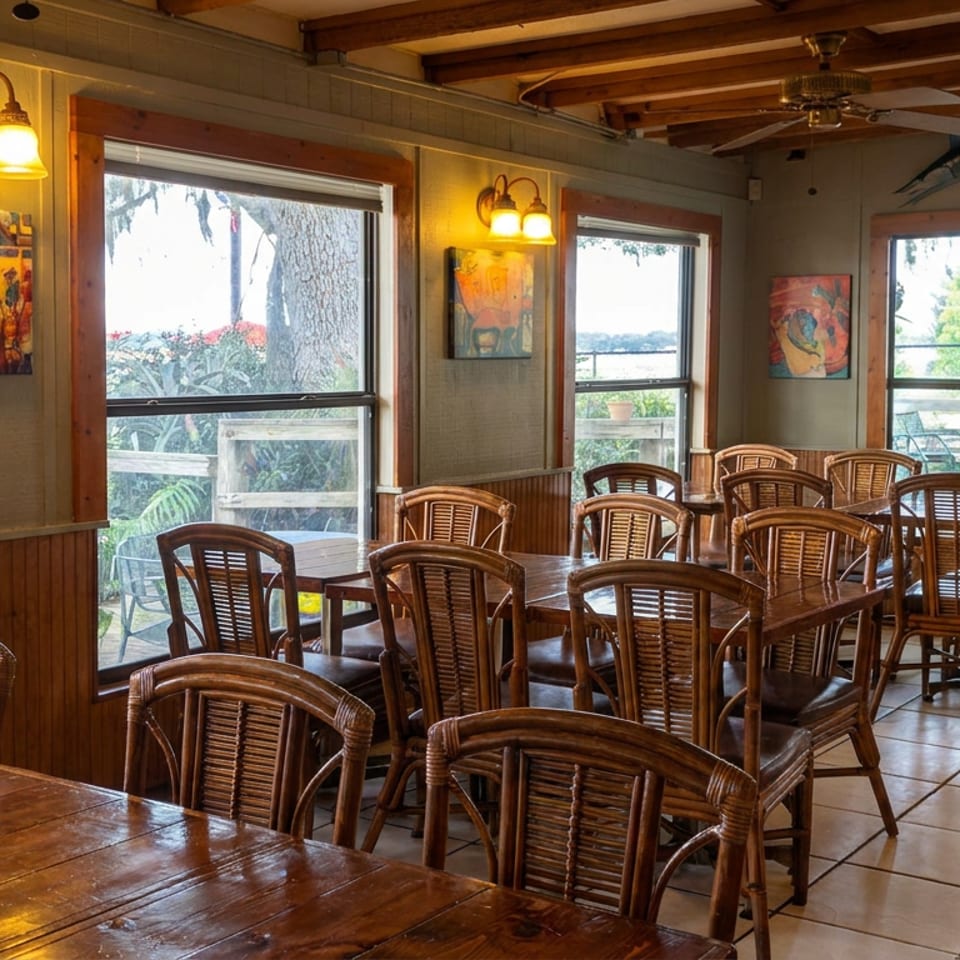 Warmly lit restaurant dining room with wooden tables, wicker chairs, artwork, ceiling fan, and windows overlooking a patio.