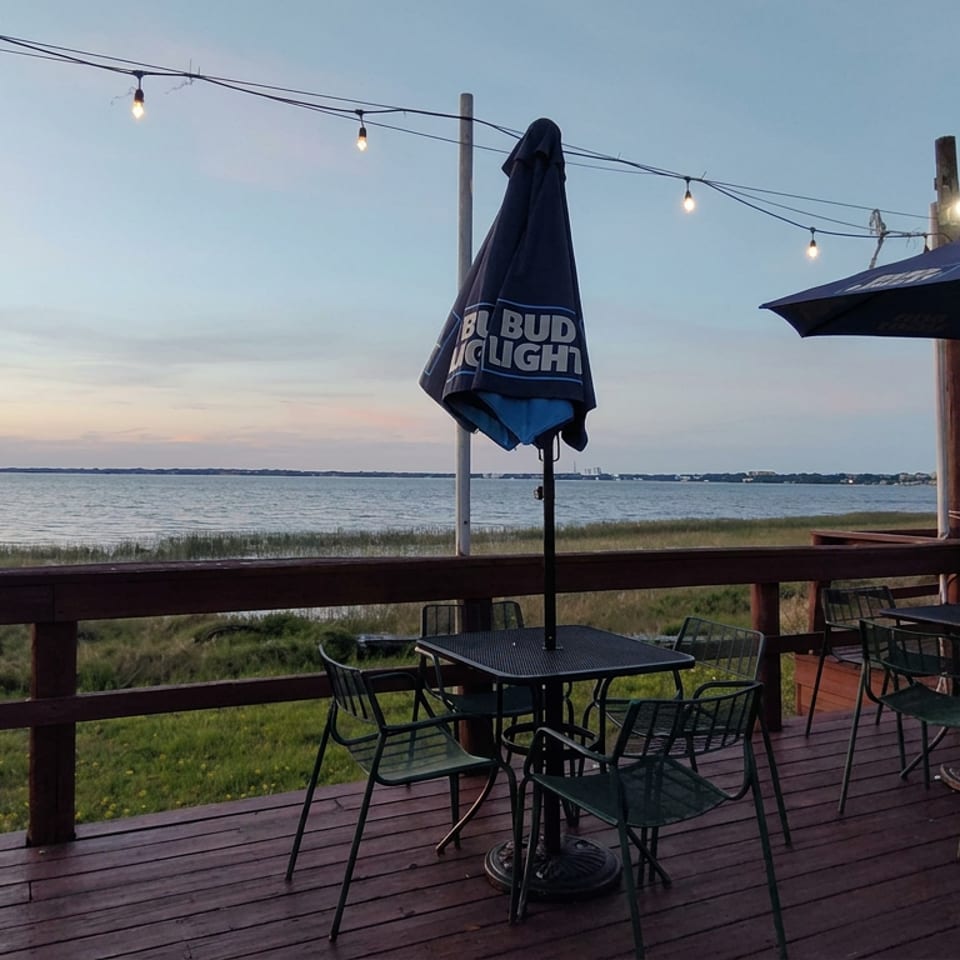 Waterfront restaurant deck with empty tables, umbrellas, string lights, and a calm bay at sunset.