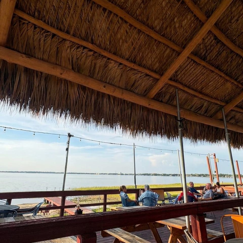Open-air waterfront restaurant with a thatched roof, string lights, wooden tables, and diners overlooking calm water.